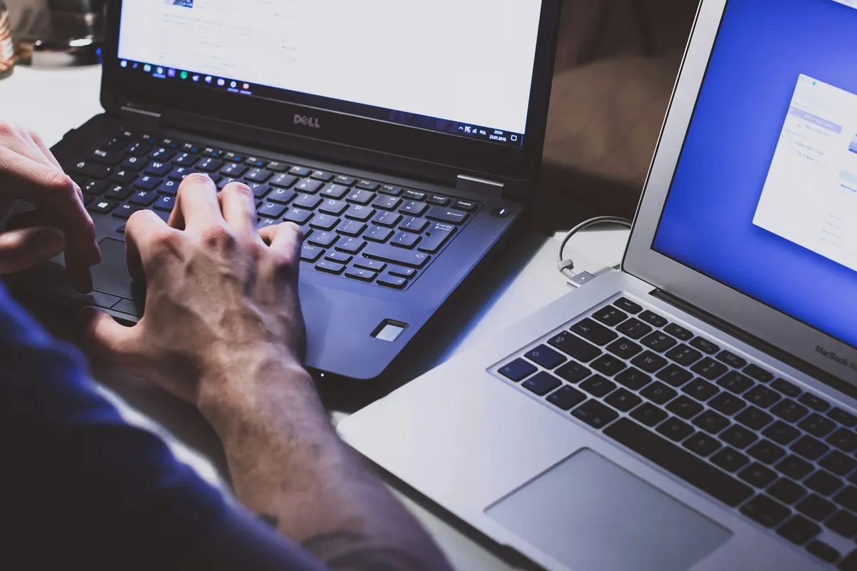 Pessoa digitando em um notebook em ambiente escuro, com outro laptop ao lado iluminado.