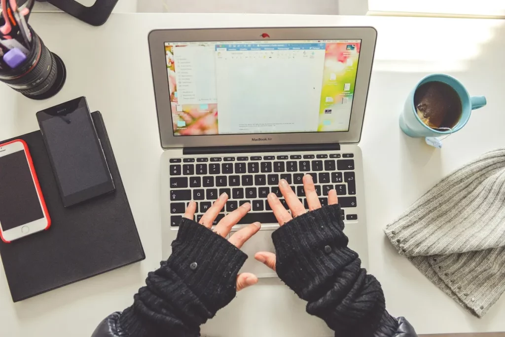 Mãos digitando em um notebook, cercado por smartphones, caderno, uma xícara de chá e um gorro sobre a mesa.