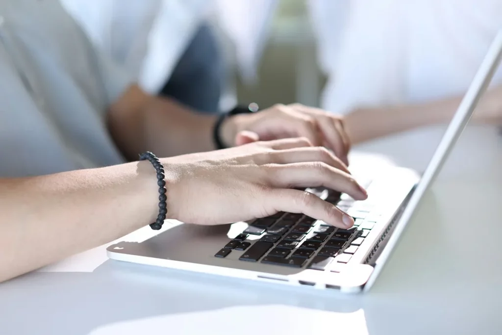 Mãos masculinas digitando em laptop prateado em ambiente iluminado, representando produtividade e trabalho em equipe.