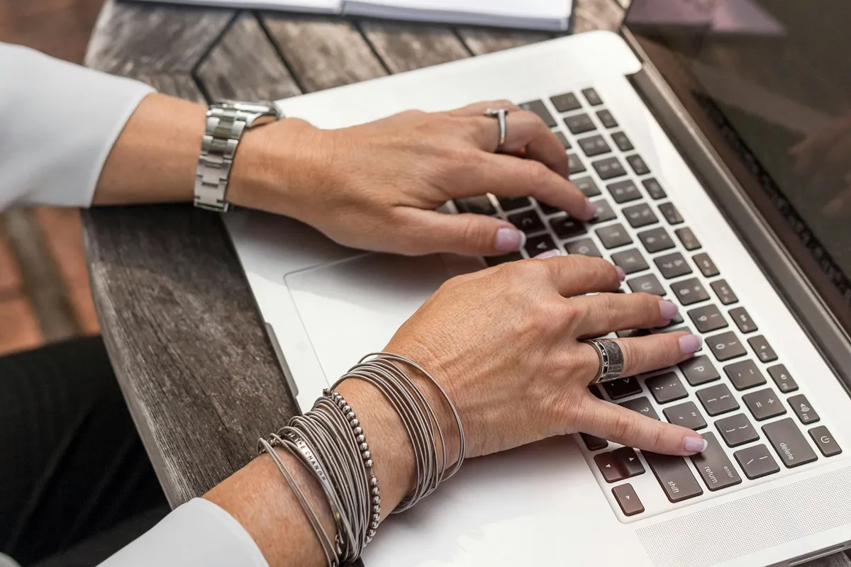 Mãos femininas usando acessórios enquanto digitam em um laptop prateado, colocado sobre uma mesa de madeira.