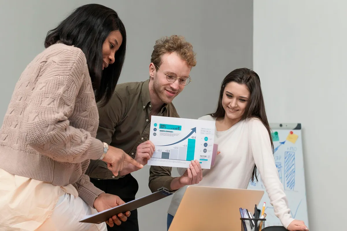 Três colegas de trabalho analisando gráficos impressos e discutindo resultados em frente a um laptop