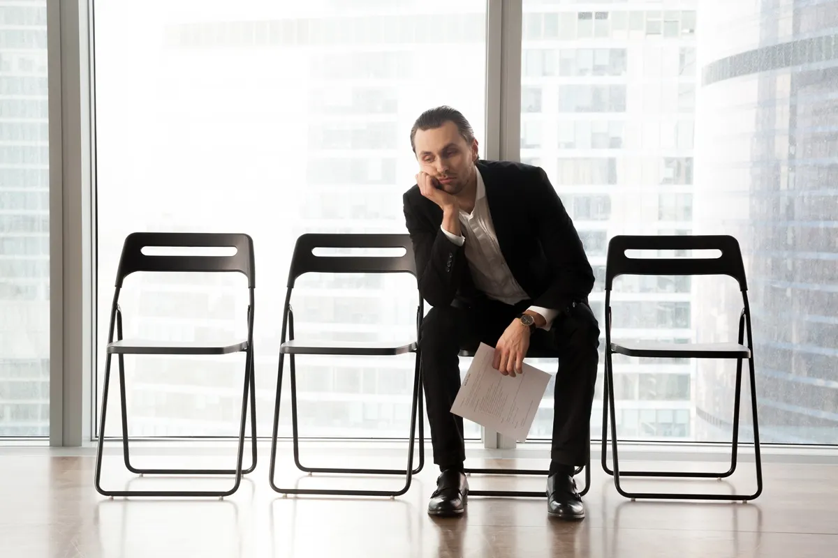 Homem desmotivado em um ambiente corporativo, sentado em uma cadeira, segurando papéis, simbolizando o impacto do absenteísmo no trabalho.