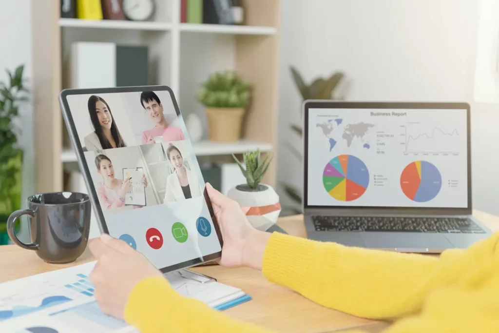 Tela de tablet exibindo uma videoconferência com quatro participantes, enquanto um notebook ao lado mostra gráficos de desempenho e relatórios visuais.