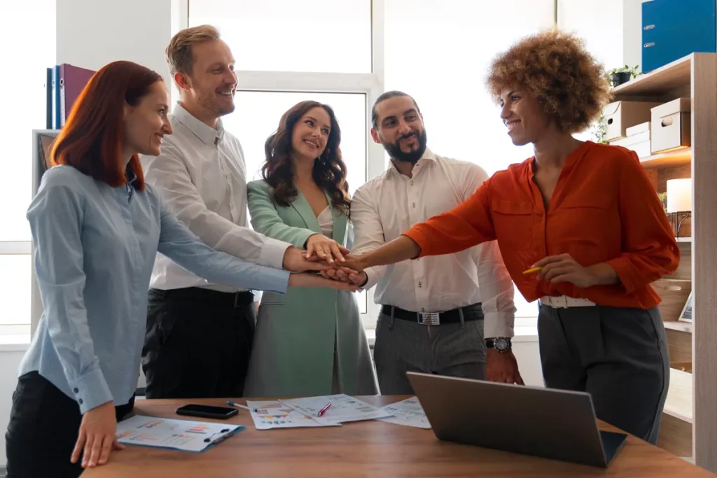 Grupo de colegas de trabalho juntando as mãos sobre uma mesa em sinal de união e trabalho em equipe, com papéis e laptop sobre a mesa.