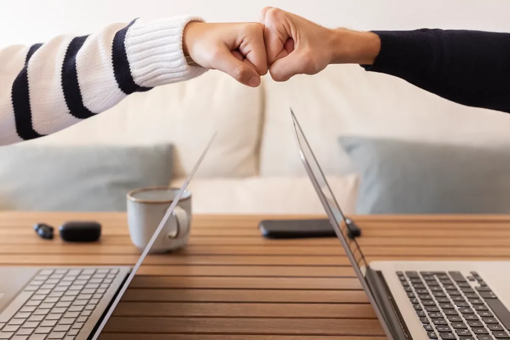 Dois colegas tocando os punhos em sinal de parceria em um ambiente de trabalho com laptops abertos sobre uma mesa de madeira