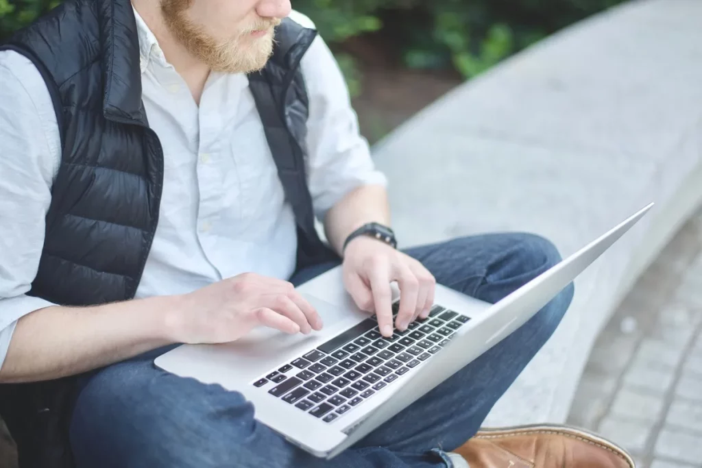 Homem digitando em um laptop enquanto está sentado ao ar livre, simbolizando flexibilidade e trabalho remoto.