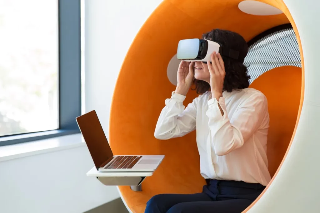 Mulher usando óculos de realidade virtual em uma cadeira moderna com laptop à frente, explorando tecnologia em ambiente corporativo.