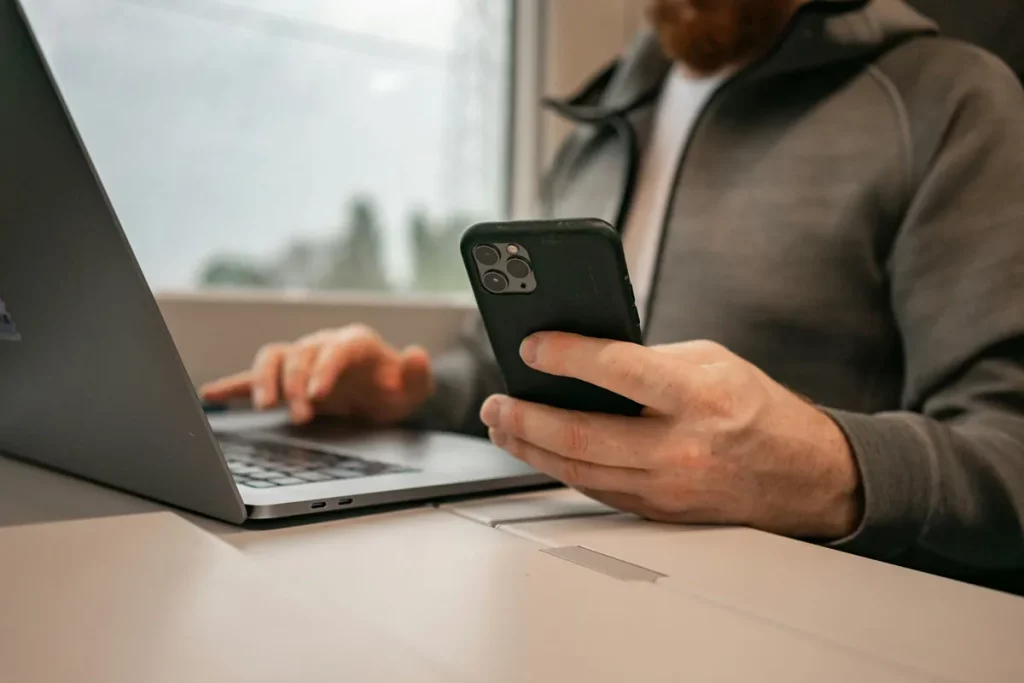 Homem segurando um smartphone enquanto usa o teclado de um laptop, simbolizando trabalho multitarefa e conectividade digital.