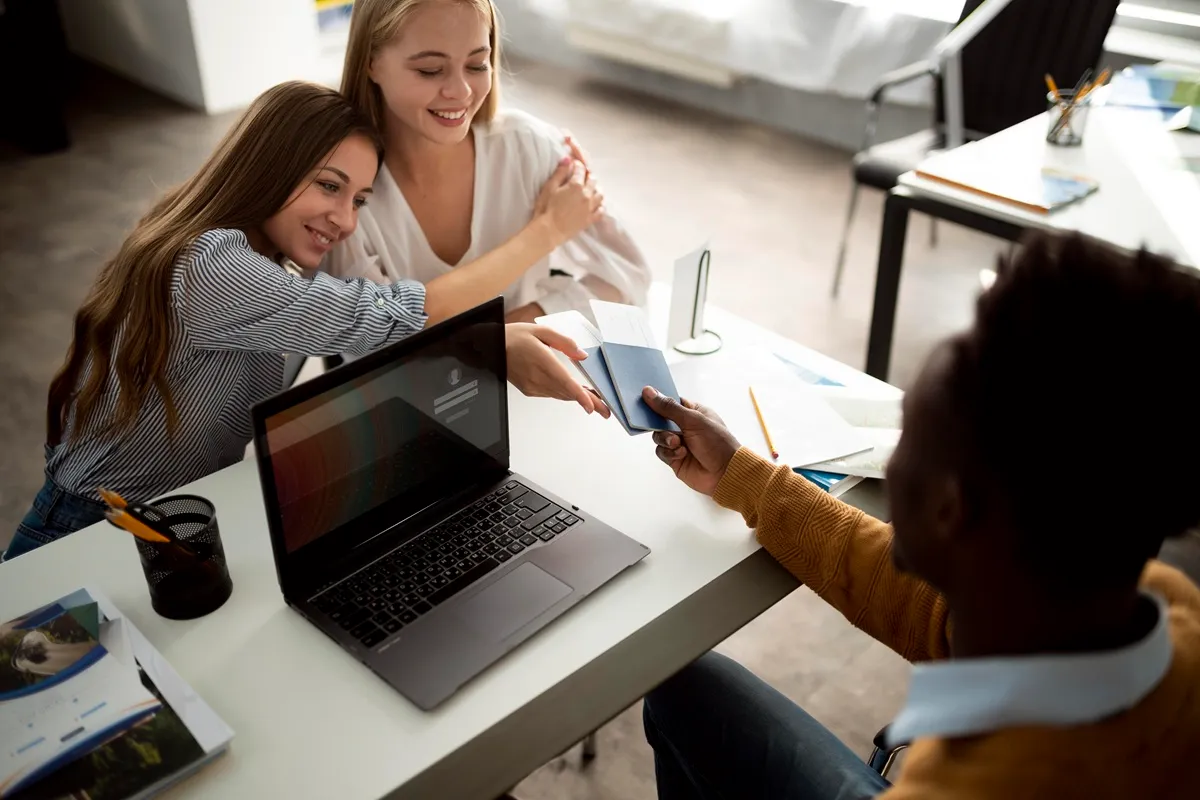 Agente de viagens entregando passaporte e documentos de viagem a duas mulheres sorridentes em um escritório com laptop aberto e materiais impressos na mesa.