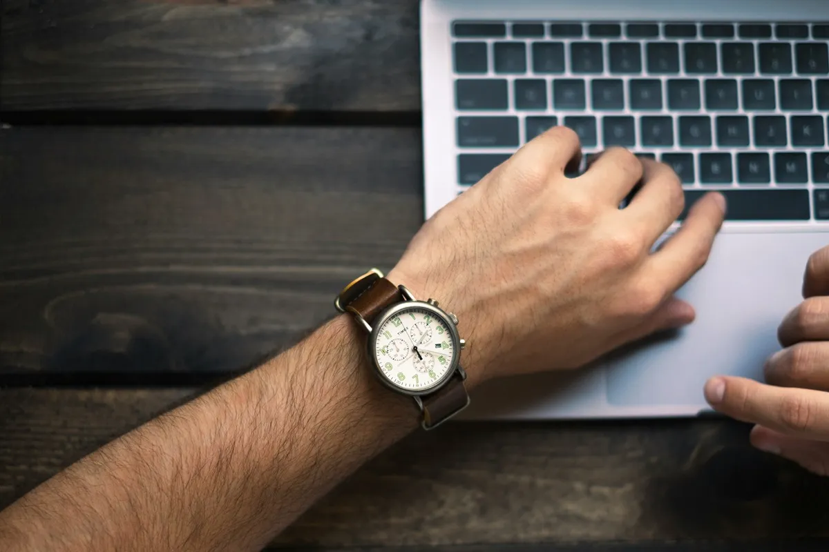 Mão masculina com relógio de pulso digitando em um laptop, simbolizando foco e gestão eficiente do tempo.