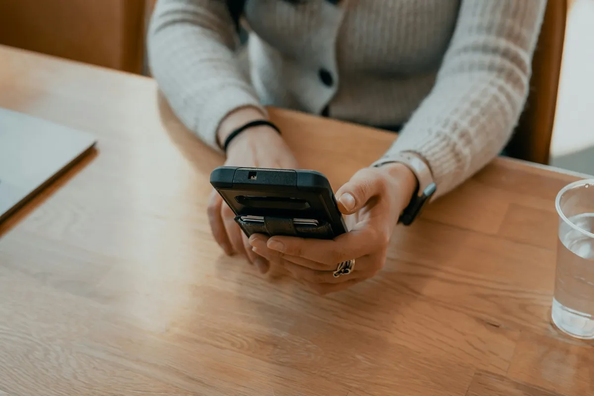 Mulher segurando um smartphone em uma mesa de madeira, utilizando o teclado virtual para comunicação ou trabalho.