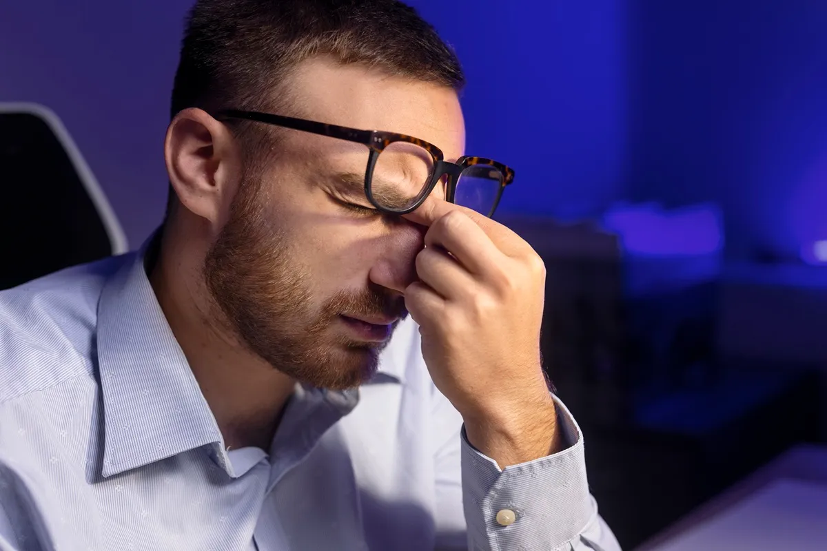 Homem de óculos pressionando a região entre os olhos, aparentando cansaço e dificuldade de concentração, em um ambiente iluminado por luz azul.