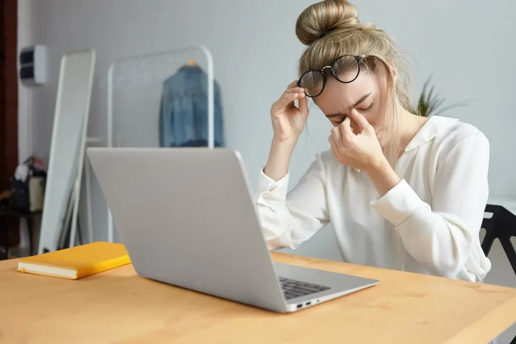 Mulher loira, com coque no cabelo, pressionando a região entre os olhos e segurando os óculos, aparentando cansaço mental e dificuldade de concentração, sentada à frente de um notebook.