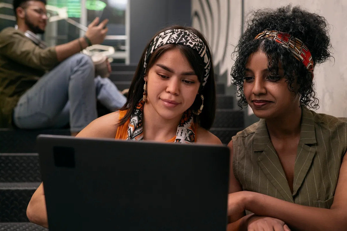 Duas mulheres jovens usando um laptop juntas, sorrindo e compartilhando conhecimento, enquanto um homem ao fundo utiliza o celular, em um ambiente moderno.