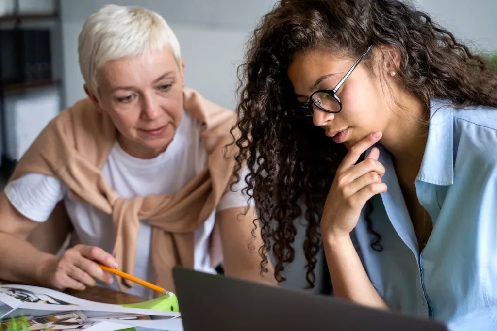 Duas mulheres de diferentes gerações trabalhando juntas em um laptop, analisando documentos e trocando ideias, representando inclusão digital e colaboração.